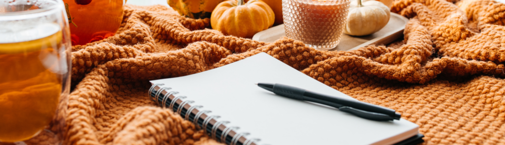 A notebook and pen on an orange knit blanket with pumpkins and candles, representing fall 2027 planning